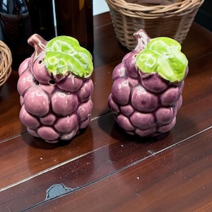 Peut inclure: Une paire de salières et poivrières en céramique en forme de grappes de raisin violettes avec une feuille vert vif sur le dessus. Chaque pièce présente une finition brillante et de petits trous sur le dessus pour le dosage, posées verticalement.
