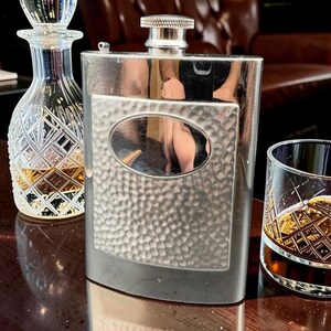 May include: A silver hip flask with a textured oval, a crystal decanter, and a patterned glass with a dark liquid. The items are on a dark wooden surface, suggesting a bar or lounge setting.