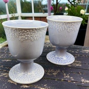 May include: Two pale blue goblets with a floral pattern around the rim. The goblets have a pedestal base and are on a distressed wooden table. The background shows a greenhouse and plants.