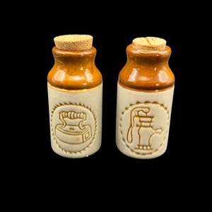 Peut inclure: Deux petites bouteilles en céramique avec des bouchons en liège, présentant un vernis bicolore crème et marron. Chaque bouteille affiche un motif circulaire en relief sur le devant, l'un représentant un fer à repasser et l'autre une pompe à eau.