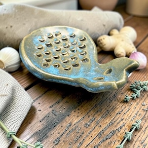 Peut inclure: Un plat à râper en céramique avec une forme de bol peu profond et arrondi, doté d'une surface texturée avec des crêtes semi-circulaires en relief. L'émail est un mélange de brun terreux et de bleu vif, avec une petite poignée saillante.