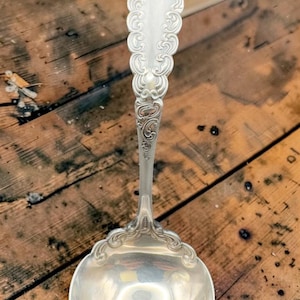 Peut inclure: Une louche en argent avec un manche orné et un bol rond. La poignée présente un motif détaillé de volutes et le bol reflète l'environnement. La louche est probablement vintage ou antique, et est un ustensile de cuisine.