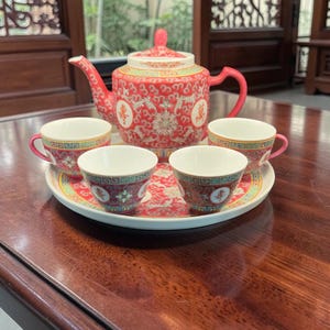 Peut inclure: Un service à thé en porcelaine rouge comprenant une théière et quatre petites tasses disposées sur un plateau rond assorti. L'ensemble est orné de motifs floraux dorés et blancs, de bordures géométriques et de caractères chinois.