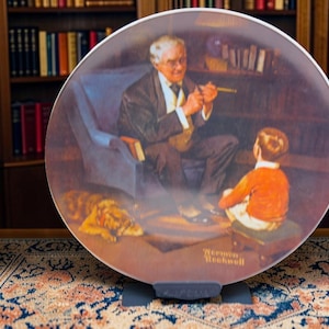 May include: A decorative plate depicting a painting of a man, a boy, and a dog in a library. The man is seated, holding a pipe, and the boy is seated on a stool. The dog is lying on the floor. The plate is displayed on a black stand.
