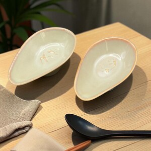 May include: Two avocado-shaped ceramic bowls with a light green glaze and a pink rim sit on a wooden table. A black spoon, a pair of chopsticks, and a folded beige napkin are also on the table. The bowls have a minimalist design.