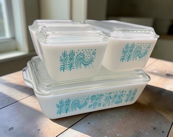 Puede incluir: Un juego de tres recipientes de almacenamiento de vidrio opalino blanco con tapas de vidrio transparente. Cada recipiente presenta un estampado gráfico en azul cerceta de gallos, tallos de trigo y motivos florales estilizados. El juego incluye un plato rectangular y dos cuadrados.