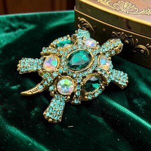 May include: A turtle-shaped brooch featuring a gold-tone metal base encrusted with small light blue rhinestones. The shell is decorated with larger oval-shaped dark green faceted stones and round iridescent stones that reflect multiple colors.