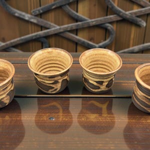 Peut inclure: Quatre tasses en céramique faites à la main avec un glaçage marron et beige. Les tasses ont une forme arrondie avec un intérieur texturé et un motif brun foncé et organique à l'extérieur. Elles sont disposées sur une surface en bois.