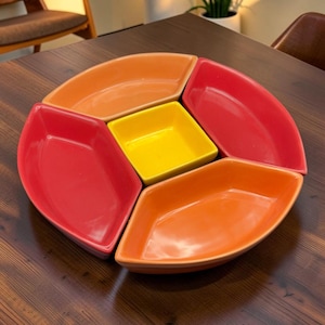 Peut inclure: Un ensemble de service en céramique composé de quatre bols en forme de feuille aux couleurs alternées orange et rouge, disposés autour d'un bol carré central jaune. Les pièces sont lisses, brillantes et présentent des bords incurvés.