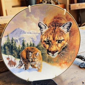 May include: A decorative plate featuring a detailed painting of two cougars in a snowy mountain landscape. The plate has a gold rim and is mounted with two clips for display. The cougars are depicted with realistic fur and intense expressions.