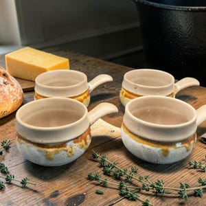 Peut inclure: Quatre bols à soupe en céramique avec poignées, dotés d'un intérieur crème et d'un extérieur blanc texturé avec une glaçure ambrée coulante. Les bols sont disposés sur une surface en bois avec un pain, du fromage et des brins de thym.