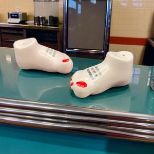 May include: Two white ceramic foot-shaped salt and pepper shakers with red painted toenails. Each shaker features black text on the top that reads, I WALKED MY FEET OFF IN WILDWOOD, N.J. The shakers have small holes on the top for dispensing seasoning.