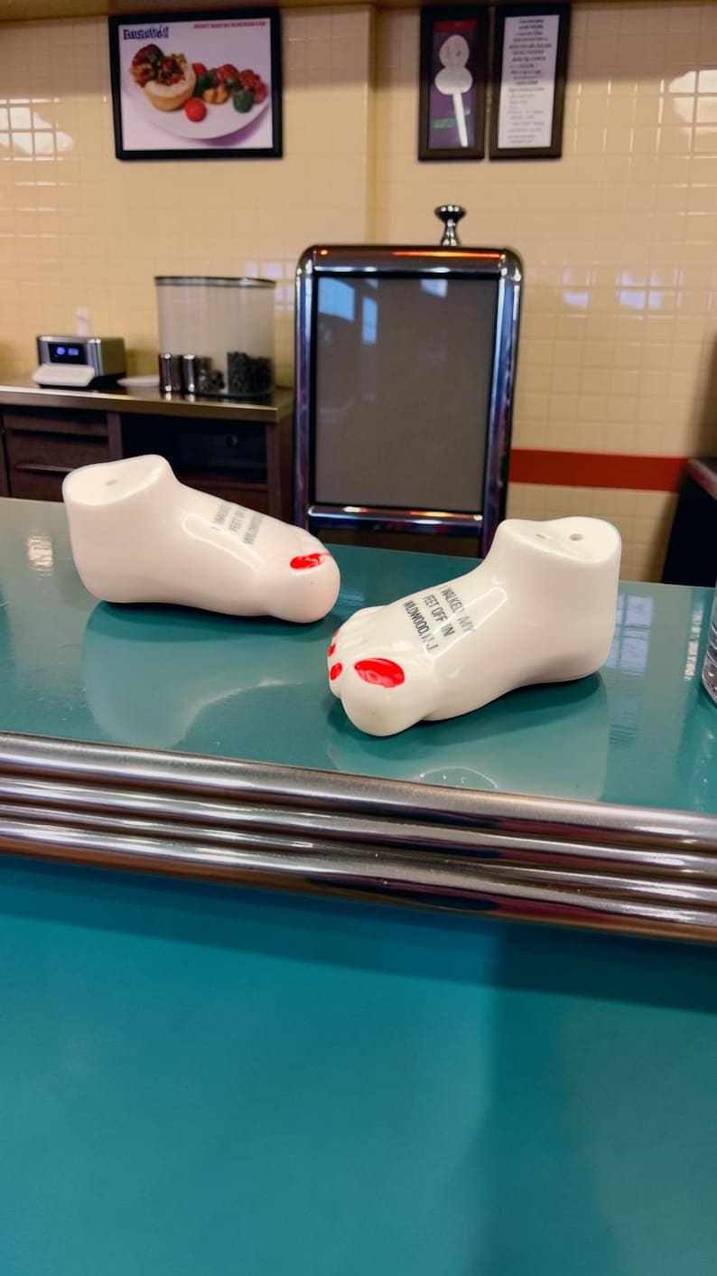 May include: Two white ceramic foot-shaped salt and pepper shakers with red painted toenails. Each shaker features black text on the top that reads, I WALKED MY FEET OFF IN WILDWOOD, N.J. The shakers have small holes on the top for dispensing seasoning.