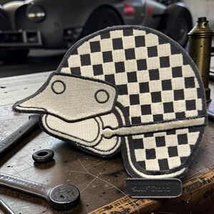 May include: A circular embroidered patch featuring a black and white checkered pattern. The design includes a stylized light gray helmet shape with two circular accents and a horizontal line. A small rectangular tag at the bottom reads Surf Stana.