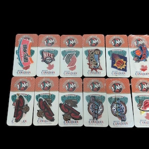 Peut inclure: Une collection de douze épinglettes avec des logos d'équipes de basket-ball NBA, chacune montée sur une carte de support individuelle orange et blanche portant le texte "JAM" et "CANADIAN". Les épinglettes présentent divers emblèmes d'équipes.