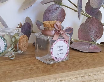Könnte beinhalten: Quadratisches Glasgefäß mit Korken und rosa Schleife. Ein florales Etikett zeigt "Baptême Louise 12 avril 2026". Das Gefäß steht auf einer Holzoberfläche mit dekorativen Blättern.