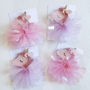 May include: Four pink unicorn hair clips with tulle and glitter. Each clip features a pink unicorn with closed eyes and a gold horn. The tulle is layered and has a shimmery finish.