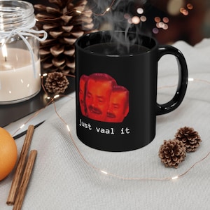 Peut inclure: Mug à café noir avec une image rouge et orange d'un visage d'homme avec le texte "just vaal it" imprimé sur le devant.