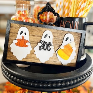 Puede incluir: Un letrero de madera con tres fantasmas blancos que sostienen una calabaza, un letrero de "Boo" y un caramelo de maíz. El letrero está en una bandeja de niveles negros con caramelo de maíz en un frasco y pajitas rayadas de naranja y blanco en una taza negra.