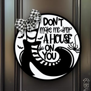 Puede incluir: Un letrero de puerta redondo en blanco y negro con un gráfico de una bota de bruja y medias a rayas con el texto "DON'T make me drop A HOUSE ON YOU". El letrero está decorado con un lazo de cuadros blanco y negro y pequeños iconos de murciélagos.