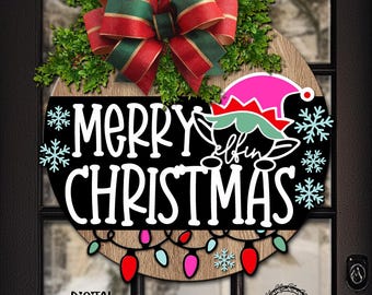 May include: A circular wooden door sign featuring the text Merry Christmas and Elfin, decorated with a green and red plaid bow, green foliage, light blue snowflakes, a pink and green elf hat, and a string of colorful Christmas light bulbs.