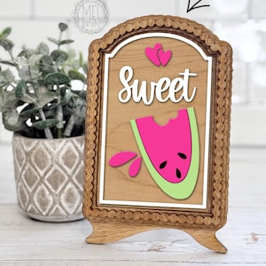 Puede incluir: Insertar letrero de madera con diseño de sandía. El letrero presenta la palabra "Sweet" en escritura blanca, una rodaja de sandía rosa con semillas negras y detalles de cáscara verde. El marco es de madera.