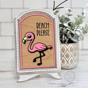 Puede incluir: Un letrero decorativo de madera con una ilustración de flamenco rosa y las palabras "BEACH PLEASE". El letrero está en un marco blanco con un fondo marrón claro. Es una descarga digital.