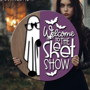 Op de afbeelding: Ronde houten bord met een cartoon spook met een bril en de tekst "Welcome to the Sheet Show", met vleermuizen en een spin. De persoon die het bord vasthoudt, draagt een zwarte jurk.