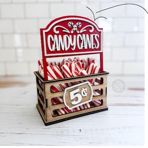 May include: A wooden candy cane stand with a red and white sign that reads "CANDY CANES". The stand is filled with red and white striped candy canes. The sign also has the text "This is an interchangeable" and "Digital file/download SVG DXF".