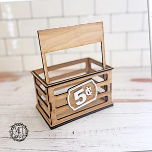 May include: A wooden crate with a sign that reads "5¢" and a blank banner. The text "This is an interchangeable" and "Digital file | download SVG DXF" are also visible. The crate is made of light brown wood.