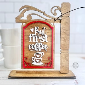 Puede incluir: Un letrero de madera con un borde rojo y el texto "But first coffee" en letras blancas. El letrero presenta una taza de café estilizada con corazones. El letrero está unido a un marco de madera.
