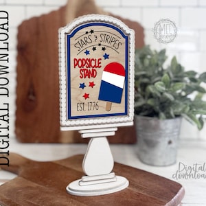 Puede incluir: Un letrero de madera de "Stars & Stripes Popsicle Stand" con un diseño de helado rojo, blanco y azul. El letrero está sobre un soporte blanco con una base decorativa. También se incluye el texto "EST. 1776".
