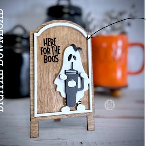May include: A wooden sign with a white ghost holding a cup that says "Here for the boos". The sign is on a wooden easel.
