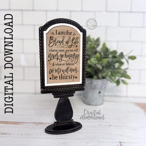 May include: A black wooden sign with the text "I am the Bread of Life" in a decorative frame. The sign is on a black stand. The background includes a plant in a metal pot and a white tile wall.