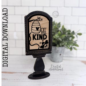 May include: A rectangular wooden sign insert featuring a black laser-cut design of a beehive, a bee, and the text BEE KIND. The insert is framed in a black scalloped border and is designed to fit into a separate stand, as indicated by the text.