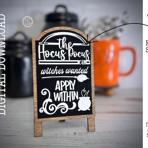 Könnte beinhalten: Ein Holzschild mit einem schwarzen Schiefertafel-Einsatz, auf dem "the Hocus Pocus Co. witches wanted APPLY WITHIN" mit einer Grafik von einem Kessel und einem Besen steht. Das Schild steht auf einer weißen Oberfläche.