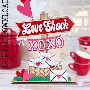 Peut inclure: Téléchargement numérique pour un projet d'artisanat sur le thème de la Saint-Valentin. Le motif présente une pancarte rouge et blanche qui dit "Love Shack" avec une bannière XOXO en dessous. La pancarte est fixée à un poteau en bois avec trois ballons en forme de cœur rose et blanc. Le motif comprend également un panier avec des enveloppes et des cœurs.