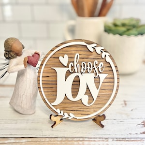Puede incluir: Un letrero redondo de madera con fondo blanco y las palabras "choose joy" en una fuente decorativa. El letrero está en un pequeño soporte de madera y tiene un corazón y hojas alrededor del texto.