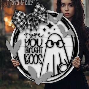 Puede incluir: Un letrero redondo de madera blanca de Halloween con un borde negro, que presenta un fantasma con gafas y el texto "Hope You Brought Boos". El letrero está decorado con murciélagos grises y un lazo a cuadros blanco y negro.