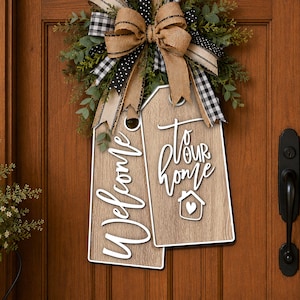 May include: A decorative door hanging featuring two light brown wood-grain tags with white text reading Welcome and to our home with a small house icon. A large bow made of burlap, black and white buffalo check, and polka dot ribbon sits above.
