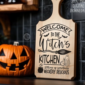 Puede incluir: Una tabla de cortar de madera con un diseño de temática de Halloween. Dice "Bienvenido a la cocina de la bruja" con una escoba, un caldero, un fantasma y murciélagos. El texto debajo dice "Preparando algo deliciosamente malvado."