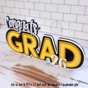 以下が含まれることがあります： 黄色、黒、白の「Congrats GRAD 26」という文字が特徴の、層状の立体的な看板。 「Congrats」は筆記体で、「GRAD」は太字のブロック体で、数字の「26」が付けられています。下部のテキストには「File is set to 15.5 x 7.5 but size as needed / scaleable file.」と書かれています。