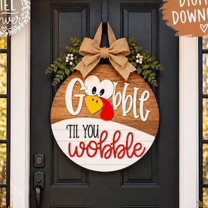 May include: A round wooden door sign featuring a turkey graphic and the text "Gobble 'til you wobble". The sign has a natural wood grain top section, a white bottom section, and is decorated with a burlap bow and green foliage at the top.