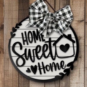 May include: A circular wooden door sign featuring a white background with horizontal black lines and the black text Home Sweet Home. The sign includes a black house outline with a heart, black leaf accents, and a black and white buffalo check bow.