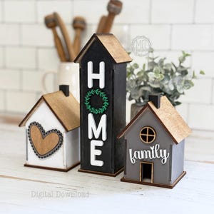 Könnte beinhalten: Drei Holzdekorationen in Form eines Hauses. Eine ist weiß mit einem Herz, eine ist grau mit dem Wort "Familie", und die höchste ist schwarz mit dem Wort "HOME" und einem Kranz. Alle haben braune Dächer.