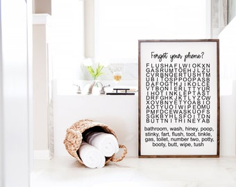 Puede incluir: Un letrero rectangular de madera con fondo blanco y marco marrón oscuro que presenta una sopa de letras titulada Forgot your phone? con una lista de palabras relacionadas con el baño como bathroom, wash, hiney, poop, stinky, fart, flush, toot.