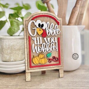 May include: A wooden tabletop sign featuring a layered design with the text "Gobble til you Wobble!" in white script. The sign includes a turkey face graphic, three yellow pumpkins, and a green leaf, all framed by a red border on a light wood background.