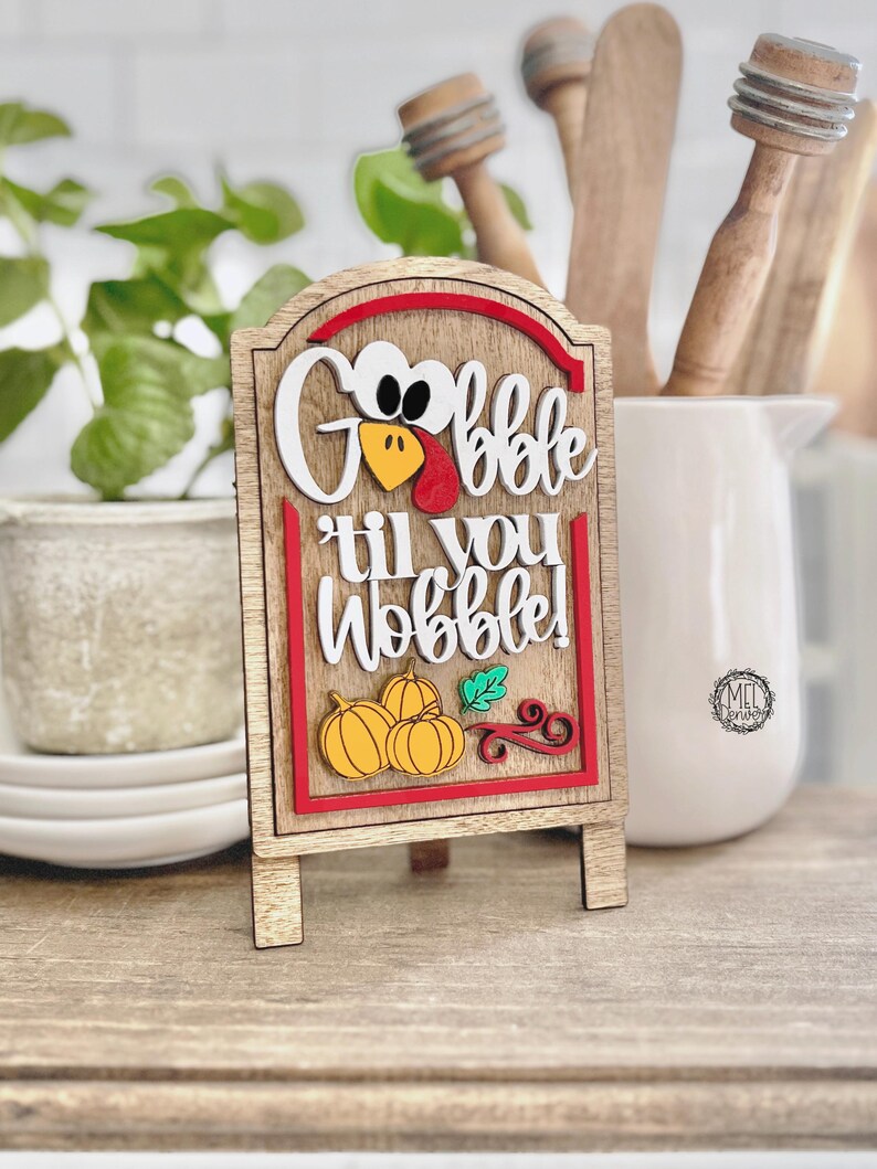 May include: A wooden tabletop sign featuring a layered design with the text "Gobble til you Wobble!" in white script. The sign includes a turkey face graphic, three yellow pumpkins, and a green leaf, all framed by a red border on a light wood background.