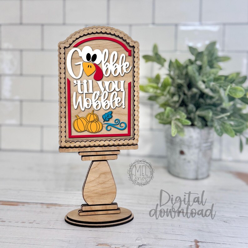 May include: A wooden tabletop sign featuring a layered design with a turkey graphic and the text "Gobble 'til you Wobble!" in white lettering. The sign includes orange pumpkin illustrations, a blue leaf, and a red border, all mounted on a pedestal base.