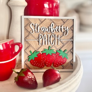 May include: Wooden sign with the words "Strawberry Patch" in white lettering. The sign has a checkered pattern and three red strawberries with green leaves.